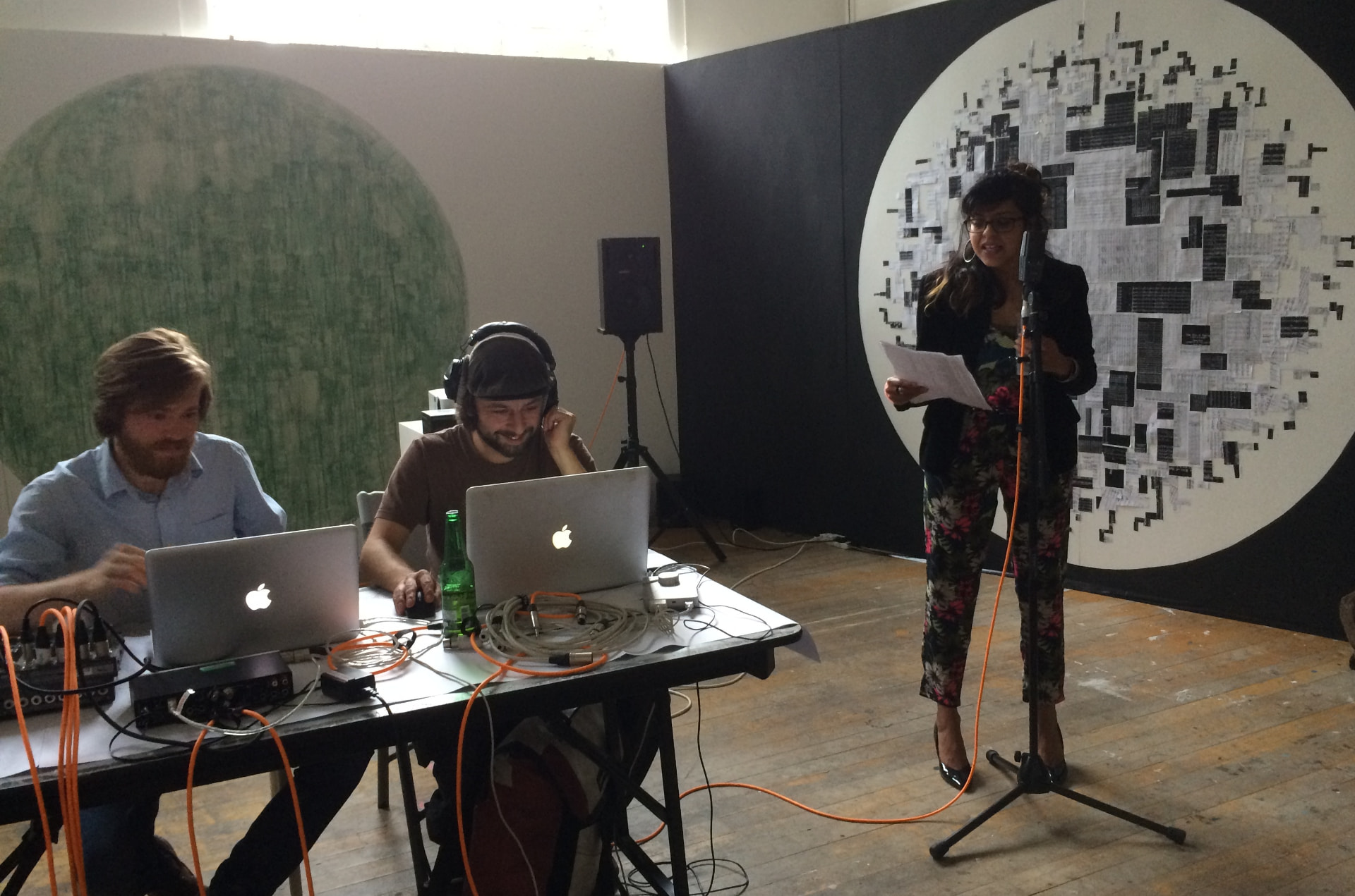 Sonia Ali performing live in front of a large circular wall work, with two sound collaborators at laptops